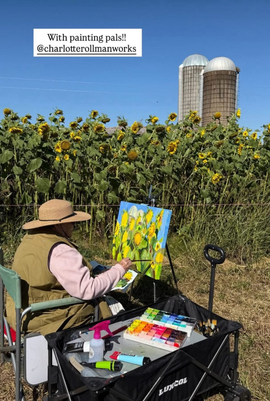 Person painting sunflowers in a field with silos in the background