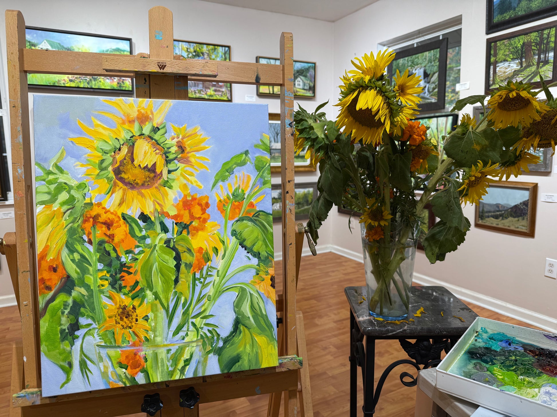 Painting of sunflowers on an easel with a vase of real sunflowers in the background. By acrylic artist Charlotte Rollman.